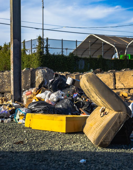 A pile of mixed household rubbish and waste materials scattered on the ground beside a low stone wall and a metal pole, including black and white plastic garbage bags, a large yellow plastic container, and an old, worn beige car seat with fabric upholstery, all indicating discarded refuse from domestic or commercial sources. The background features a green hedge, utility poles with overhead cables, and a partially visible roofed structure, suggesting an outdoor area such as a roadside or an informal dumping site. The scene is illuminated by natural daylight, emphasizing the contrast between the waste and the environment, illustrating the kind of unmanaged rubbish that private disposal services like Rubbish Removal Hounslow may handle to prevent illegal dumping and support on-site waste clearance, aligning with the theme of avoiding hidden rubbish charges and unsightly waste accumulation in the local area.