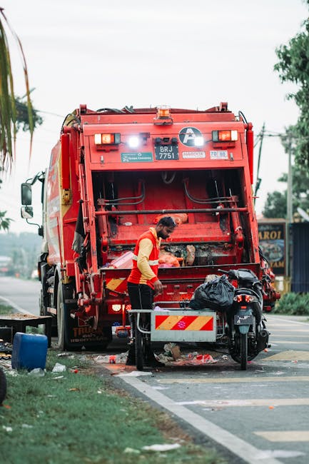 A worker wearing a yellow high-visibility vest and black trousers is operating a red rubbish collection truck parked on the side of a street, with the rear compactor open and some debris visible beneath it. The truck has various labels and reflective strips, and the worker is seen handling black rubbish bags placed nearby, including one on a small metal platform. The setting appears to be an urban or suburban street, with trees, utility poles, and a partial view of a building or shopfront in the background. The scene is illuminated by natural daylight, highlighting the metallic and plastic textures of the truck and waste bags, and reflecting typical conditions during on-site waste collection activities. The environment suggests an independent waste removal process, aligning with private rubbish handling services that may serve domestic or commercial clients seeking alternative rubbish disposal options, as provided by companies like Rubbish Removal Hounslow.