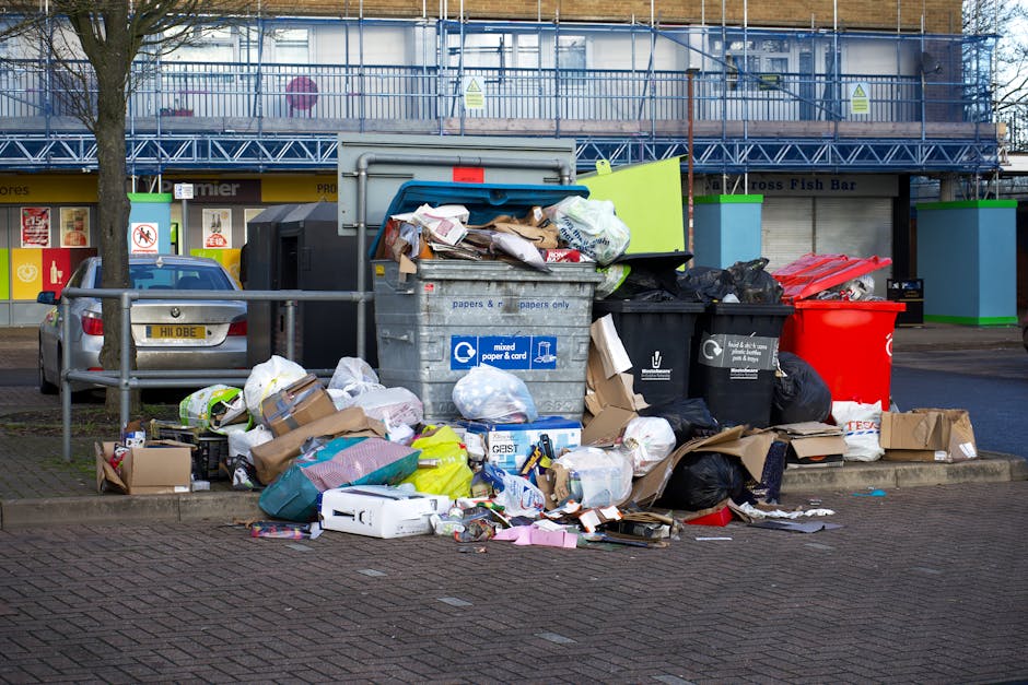 A large pile of mixed waste and rubbish bins situated on a paved urban street corner, with various discarded items including cardboard boxes, plastic bags, paper waste, and flattened packaging overflowing from open and closed bins. The waste is scattered on the ground around the bins, which are positioned near a metal railing and adjacent to a parked silver car with a UK registration plate. In the background, there is a commercial building with shopfronts and signs, along with a blue scaffolding structure covering part of the upper facade. The scene appears to be in an area where waste collection or disposal is taking place, possibly indicating a period of delayed rubbish removal or an on-site collection. The environment is well-lit with natural daylight, and the overall setting emphasizes the importance of proper rubbish management, as provided by services like Rubbish Removal Hounslow.