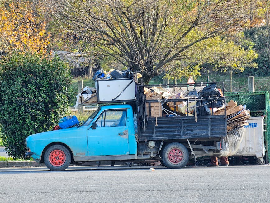 A worker wearing a yellow high-visibility vest and black trousers is operating a red rubbish collection truck parked on the side of a street, with the rear compactor open and some debris visible beneath it. The truck has various labels and reflective strips, and the worker is seen handling black rubbish bags placed nearby, including one on a small metal platform. The setting appears to be an urban or suburban street, with trees, utility poles, and a partial view of a building or shopfront in the background. The scene is illuminated by natural daylight, highlighting the metallic and plastic textures of the truck and waste bags, and reflecting typical conditions during on-site waste collection activities. The environment suggests an independent waste removal process, aligning with private rubbish handling services that may serve domestic or commercial clients seeking alternative rubbish disposal options, as provided by companies like Rubbish Removal Hounslow.