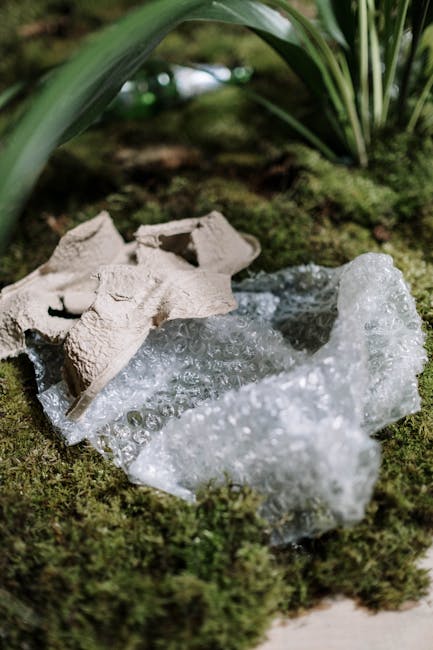 A close-up image showing broken pieces of white polystyrene foam and crumpled cardboard on a surface covered with green moss and small plants. The foam fragments are irregular in shape with a textured surface that reflects light, while the cardboard remains partially intact with fluted layers visible. In the background, there are broad green leaves and blurred natural foliage, indicating an outdoor garden environment. The scene is illuminated by natural daylight, highlighting the contrast between the synthetic rubbish and the organic surroundings. This photograph exemplifies the type of waste material managed during private rubbish clearance services, such as those provided by Rubbish Removal Hounslow, who frequently handle garden-related waste and debris in environmentally sensitive locations.