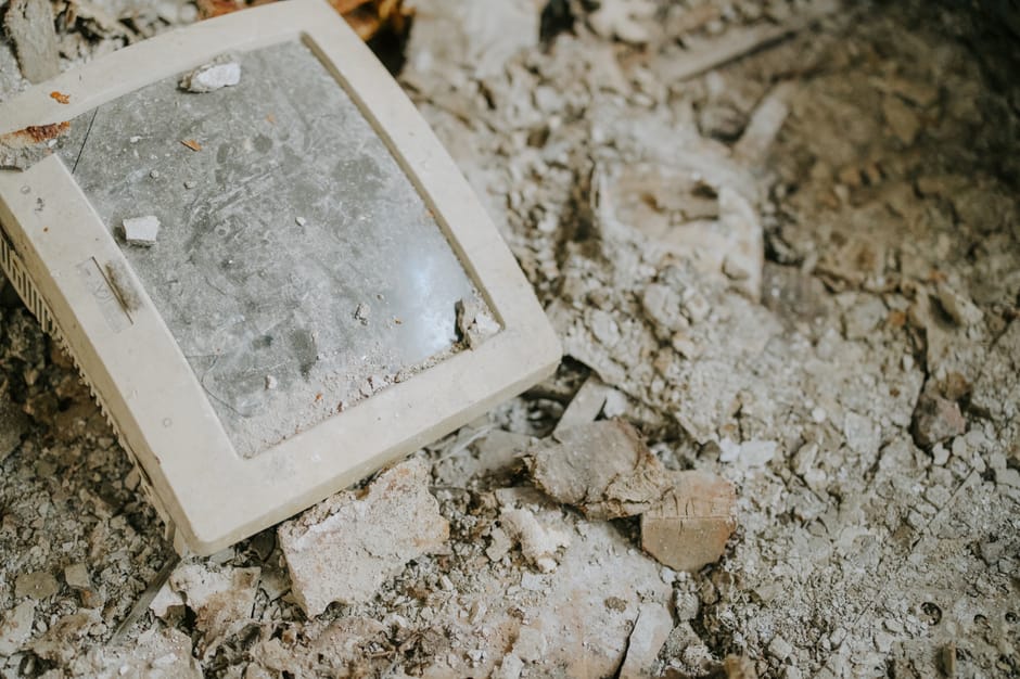 A close-up photograph of a dirty, dust-covered electronic device, possibly an old CCTV monitor or small television, lying on a surface of loose rubble, broken bricks, and dust. The device has a rectangular shape with a white plastic frame surrounding a glass screen that is covered in grime, dust, and small debris. The screen appears scratched and obscured, with scattered dirt particles and fragments of plaster or cement adhered to its surface. The surrounding environment consists of rough, uneven ground composed of broken bricks, powdered cement, and dirt, indicating a construction or demolition site. The lighting is natural, casting soft shadows across the rubble, emphasizing the textures of the debris and the aged condition of the device. This visual scene exemplifies an instance where discarded or outdated electronics lie amidst debris, reflecting aspects of waste management, disposal, or decluttering efforts that may be associated with professional rubbish removal services like Rubbish Removal Hounslow, who facilitate the clearing of such unwanted items from various environments.