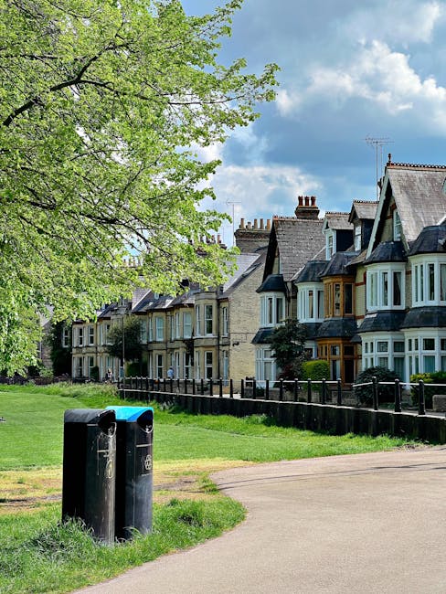 A row of traditional terraced houses with brick facades, bay windows, and pitched roofs, situated in a residential street under a partly cloudy sky. In the foreground, two black rubbish bins with blue lids are placed on a well-maintained grassy verge near a paved pathway. A large tree with bright green leaves extends its branches over the scene, providing shade and adding natural detail to the urban environment. A black wooden fence runs along the front of the houses, separating the gardens from the street. The overall scene depicts a calm suburban area with clear, natural lighting, representing typical elements of local residential waste management and private rubbish disposal, consistent with services offered by companies like Rubbish Removal Hounslow.