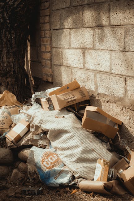 An outdoor scene depicting a small pile of mixed waste materials located against a concrete block wall. The pile includes flattened cardboard boxes, plastic bags, and packaging, with some boxes partially open or crumpled. The cardboard appears to be of standard brown kraft material, with visible corrugation edges and printed labels. The plastic bags are semi-transparent and beige in color, blending with the earthy ground surface. The waste furthest from the wall extends onto a dirt or gravel surface, with small stones scattered around. To the left of the waste pile, there is a tree trunk with dark, rough bark, and the background features a weathered wall made of large, gray concrete blocks showing slight discoloration and surface texture. The scene is lit by natural daylight, creating soft shadows that highlight the irregular textures of the waste and the surrounding environment. This imagery relates to private waste collection or on-site clearance services provided by companies like Rubbish Removal Hounslow, helping to illustrate the process of managing waste outside regular refuse collection. Overall, the scene emphasizes visual aspects of rubbish accumulation in an outdoor urban or semi-urban setting, relevant to alternative waste handling methods.