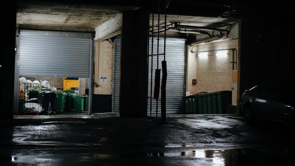 The image depicts the interior of a dimly lit underground parking garage or loading bay area, featuring two separate sections with roller shutter doors, one partially open revealing a storage space with waste collection bins and miscellaneous items, and the other closed. To the left, behind the partially open shutter, there are multiple green recycling bins and black waste containers positioned on a concrete floor that appears slightly wet, reflecting the limited light. The surrounding walls are constructed from concrete blocks painted in a light beige tone, with exposed pipes and electrical conduits running along the ceiling and walls. In the right section, a dark-colored car is partially visible, parked adjacent to the closed shutter, with its sleek surface reflecting the minimal lighting. Two vertical stainless steel poles with red padding are situated between the sections, serving as protective bollards. The scene's overall atmosphere suggests an environment suitable for private waste collection or on-site rubbish management, consistent with the services provided by Rubbish Removal Hounslow, reinforcing the context of independent rubbish handling outside of council collection points.