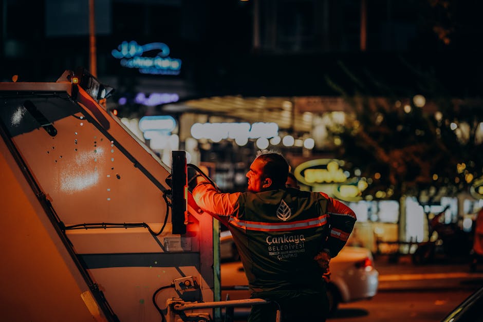 A sanitation worker wearing a high-visibility jacket with reflective stripes, labeled with the words 'Çankaya Belediyeesi,' is depicted at night operating a waste collection vehicle. The worker, seen from behind, is reaching up to the side of the vehicle, which has a smooth, orange surface with a slightly wet appearance. The background features illuminated signs and blurred city lights, indicating an urban environment with commercial establishments. The scene is lit by ambient street lighting, casting a warm glow on the worker and the vehicle. This visual context reflects an on-site waste collection activity, possibly an independent rubbish removal service, with the worker preparing or managing rubbish collection operations in a public area.