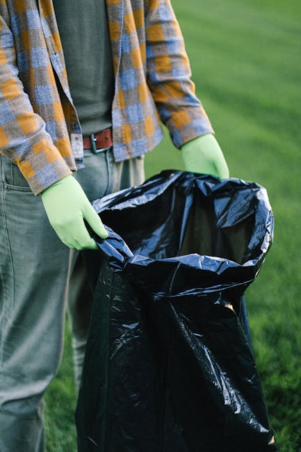 A person wearing a checkered shirt in shades of yellow, grey, and blue, with green gloves, is holding open a large black trash bag lined with reflective silver material. The individual’s grey pants and brown belt are visible, and they are standing outdoors on a grassy area with a blurred green background, likely in a park or garden. The scene suggests an active waste collection or rubbish removal activity, possibly involving urban or garden clearance, with the empty trash bag ready to be filled with rubbish. This image illustrates an aspect of professional waste management as performed by Rubbish Removal Hounslow, emphasizing readiness for on-site clearance and private disposal tasks with attention to environmental cleanliness and responsible waste handling.
