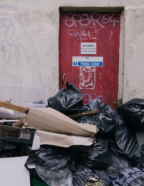 A pile of black rubbish bags and discarded cardboard and plastic waste is stacked in front of a red metal door with graffiti tags and multiple stickers, including one indicating 'No Money' and another instructing to 'Keep Clear.' The bags, some torn, contain various waste materials and are placed on a pavement area adjacent to an exterior building wall that has a rough, light-colored textured surface. The door appears weathered, with patches of faded paint and graffiti. The scene suggests a clearance or collection of waste outside a premises, potentially related to independent waste disposal, with Rubbish Removal Hounslow providing private rubbish removal services in the local area for non-council waste management solutions.