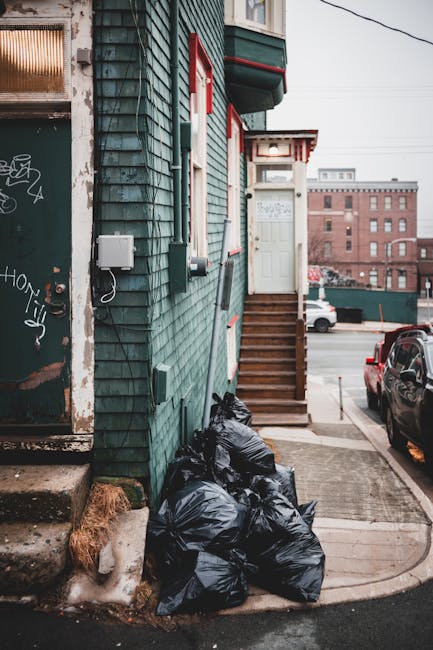 A small urban building facade with green wooden cladding, featuring a white door accessed by a short flight of wooden stairs with a small landing. To the left, there is a graffiti-covered metal door with a weathered surface, showing rust and peeling paint. Several black garbage bags are stacked on the concrete pavement at the base of the building, close to a patch of frost and dried grass. The surrounding environment includes parked cars on a street, with a larger brick building visible in the background under an overcast sky. An electrical box and wiring are attached to the building's exterior wall, and a white utility meter is installed nearby. This scene depicts a typical urban setting where private waste collection services, such as the ones offered by Rubbish Removal Hounslow, provide alternative rubbish disposal methods for residents, especially where access for larger collection vehicles might be limited or challenging.