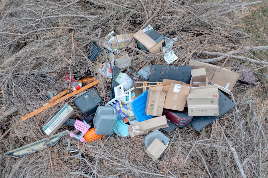 A pile of black rubbish bags and discarded cardboard and plastic waste is stacked in front of a red metal door with graffiti tags and multiple stickers, including one indicating 'No Money' and another instructing to 'Keep Clear.' The bags, some torn, contain various waste materials and are placed on a pavement area adjacent to an exterior building wall that has a rough, light-colored textured surface. The door appears weathered, with patches of faded paint and graffiti. The scene suggests a clearance or collection of waste outside a premises, potentially related to independent waste disposal, with Rubbish Removal Hounslow providing private rubbish removal services in the local area for non-council waste management solutions.