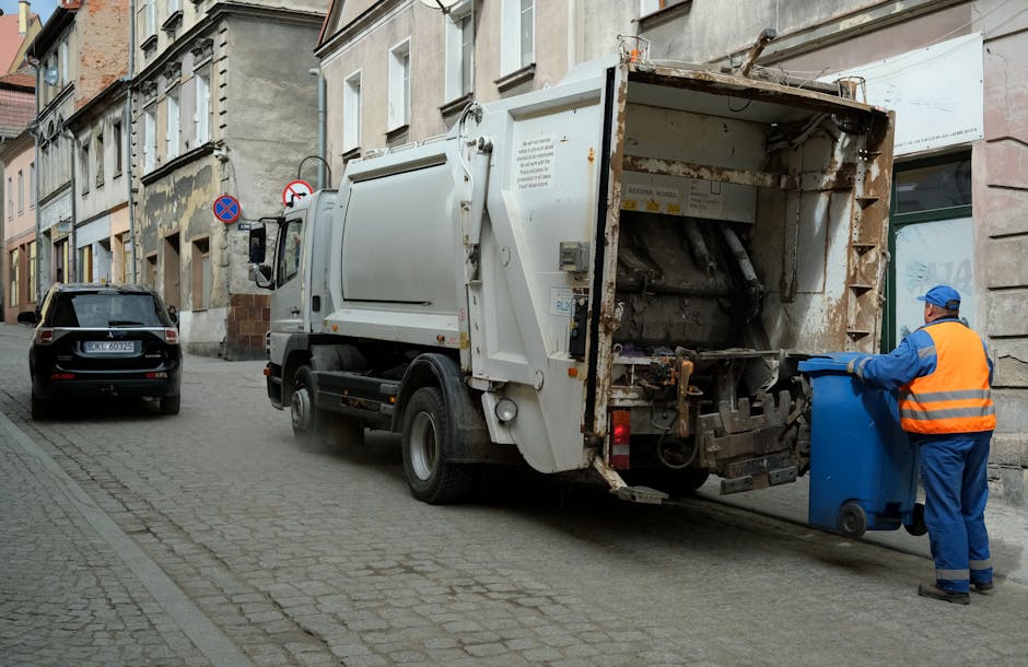 A street scene showing a white waste collection truck with signs of rust and operational equipment at the rear, positioned parallel to a curb on a paved, cobblestone street lined with multi-story buildings. A worker dressed in a blue uniform with an orange high-visibility vest, wearing a blue cap and gloves, is seen standing beside the truck, pushing a blue wheeled bin towards the loading mechanism. The background includes a parked black car with a visible license plate, and the buildings exhibit mixed architectural styles with stone and brick façades, some with windows and signage. The environment appears to be in an urban area with natural lighting, suggesting daytime, and the scene depicts typical private waste collection activity, supporting alternatives to municipal rubbish disposal services.
