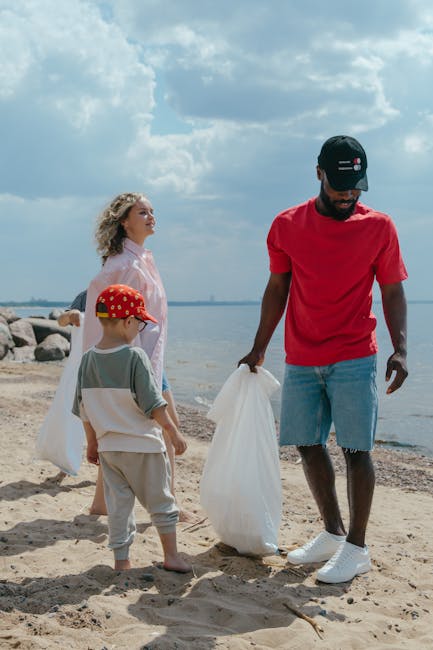 A group of three people, consisting of a man, a woman, and a young boy, are engaged in a beach clean-up by the shoreline under a partly cloudy sky. The man, positioned on the right, is wearing a black cap, a bright red T-shirt, and faded denim shorts, and is holding a white trash bag with both hands, appearing to gather litter from the sandy beach. The woman, on the left, has curly blonde hair and is dressed in a light pink raincoat, standing slightly behind the man, observing the activity while holding a bag. The young boy, in front of the woman, is wearing a red cap with a yellow pattern, a light grey T-shirt, and grey shorts, and is attentively looking at the man as he participates in the cleanup. The background features large rocks along the water's edge, calm water extending to the horizon, and a sky filled with a mix of bright and dark clouds, creating a natural environment that reflects responsible waste management and environmental care, similar to the services provided by Rubbish Removal Hounslow for private rubbish collection and on-site clearance efforts.