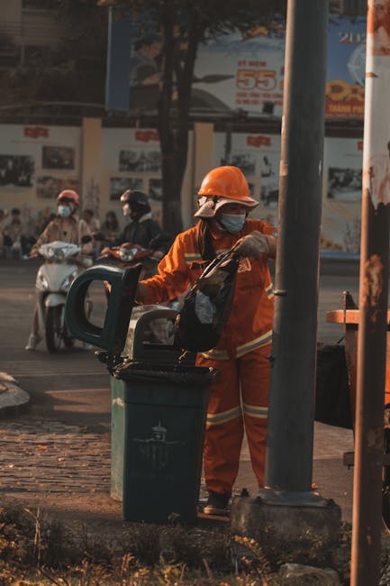 A street scene showing a white waste collection truck with signs of rust and operational equipment at the rear, positioned parallel to a curb on a paved, cobblestone street lined with multi-story buildings. A worker dressed in a blue uniform with an orange high-visibility vest, wearing a blue cap and gloves, is seen standing beside the truck, pushing a blue wheeled bin towards the loading mechanism. The background includes a parked black car with a visible license plate, and the buildings exhibit mixed architectural styles with stone and brick façades, some with windows and signage. The environment appears to be in an urban area with natural lighting, suggesting daytime, and the scene depicts typical private waste collection activity, supporting alternatives to municipal rubbish disposal services.