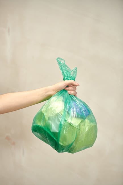 A person's hand is shown holding a green plastic rubbish bag by its twisted top, with the bag appearing full and slightly translucent, revealing some light-colored contents inside. The bag is made of thin, flexible plastic material with a glossy finish, reflecting a minimal amount of light. The hand, visible from the wrist to the fingers, is positioned on the left side of the image, gripping the bag firmly. The background is plain, neutral-colored wall with no additional objects or environmental elements visible, emphasizing the plastic bag as the main focus. This scene relates to waste management and rubbish collection services offered by companies such as Rubbish Removal Hounslow, conveying a clear image of the typical waste disposal process using plastic refuse bags. The lighting is soft and even, creating a neutral tone suitable for informational and service-related content about private rubbish removal or alternative waste handling methods in the context of Hounslow West station area.